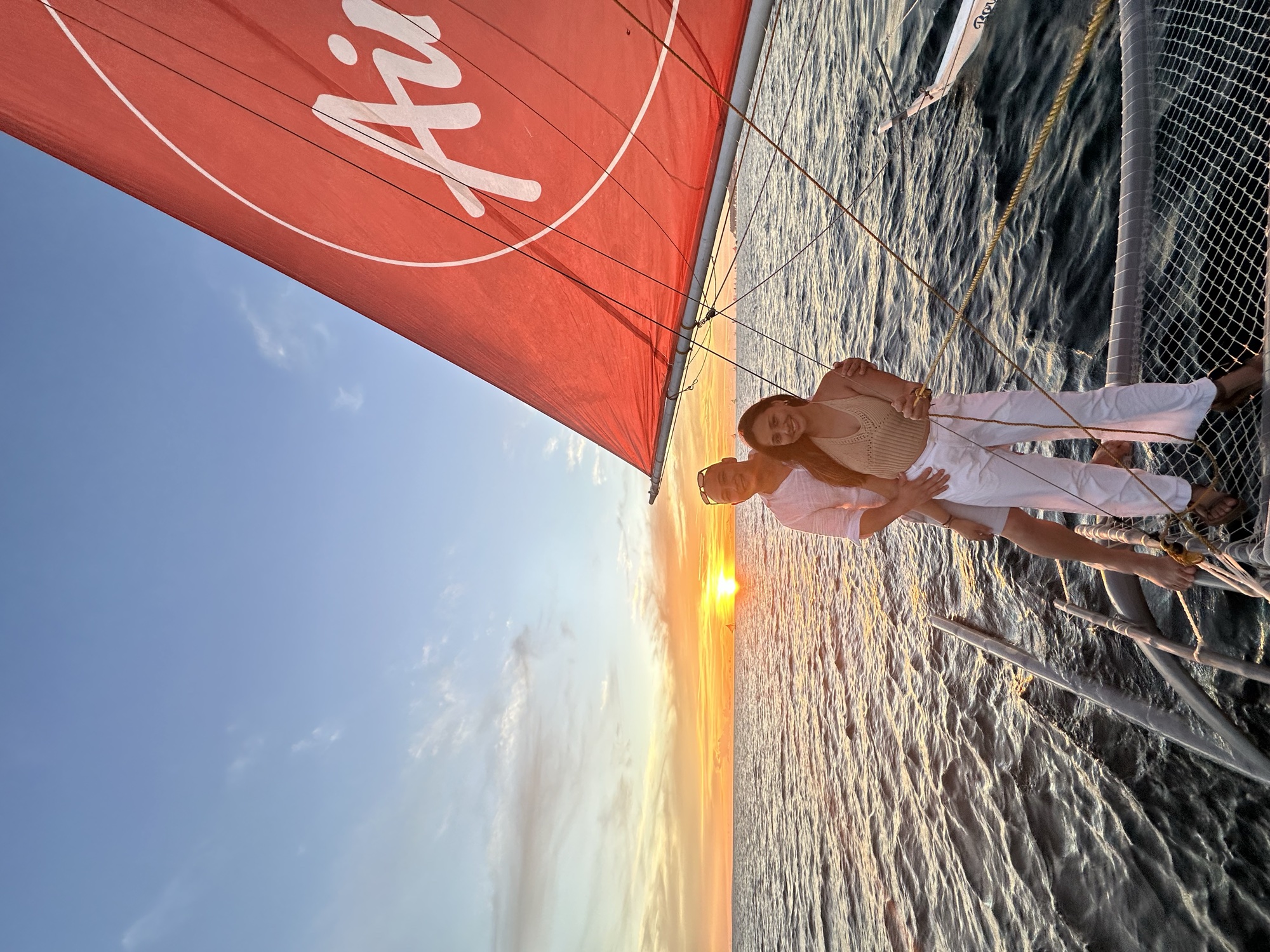 David and Chai standing together at the bow of a sailboat at sunset, the red sail above them.